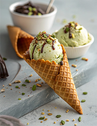 Pistachio ice cream scoops in a waffle cone, drizzled with chocolate and topped with crushed nuts.