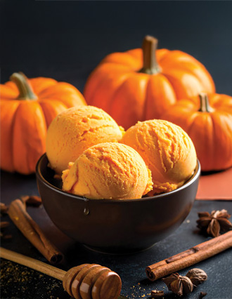 Three scoops of pumpkin ice cream in a bowl, with pumpkins, cinnamon sticks, and honey in the background.